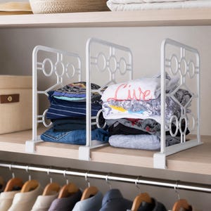 May include: A white metal shelf divider with a geometric circular pattern, featuring two vertical panels that clip onto a wooden shelf to organize stacks of folded clothing, including a white shirt with the red text Live printed on the front.