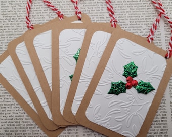May include: Set of six rectangular gift tags. Each tag has a brown border and a white embossed center with a floral design and a green and red holly accent. The tags are strung with red and white twine.