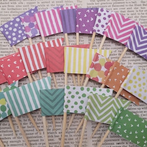 May include: A selection of miniature paper flags on wooden skewers, displaying diverse patterns such as polka dots, stripes, and chevrons. The flags are in various colours, including pink, green, and orange. The backdrop is a vintage newspaper.