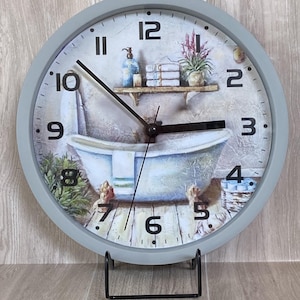 May include: A round wall clock with a light gray frame and a printed face featuring a bathroom scene with a clawfoot tub, a shelf with towels, a plant, and black numerals. The clock has black hour, minute, and second hands centered on the dial.