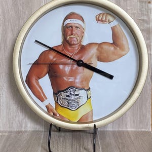 May include: Round clock with a gold-colored frame featuring a picture of a wrestler. The wrestler is wearing a white headband, yellow trunks, and a championship belt. The clock has black hands and sits on a black metal stand.