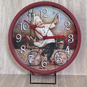 May include: A burgundy-colored wall clock with a whimsical illustration of a chef riding a bicycle. The clock face features large white numerals and red hands. The chef is depicted with a handlebar mustache and a white chef's hat.
