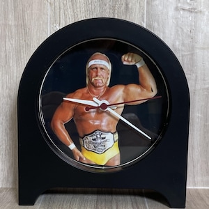 May include: A black, arched desk clock featuring a picture of a muscular man in yellow trunks and a championship belt. The clock hands are white with red accents. The clock is approximately 6 inches tall.