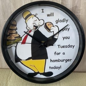 May include: A round wall clock with a black frame and a white face featuring a cartoon character wearing a yellow hat, black jacket, and yellow trousers. The character holds a hamburger and a tray of burgers with the text I will gladly pay you Tuesday for a hamburger today!
