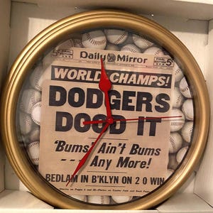 May include: A round gold-colored clock with a glass face. The clock hands are red. The clock face features a newspaper clipping with the headline "DODGERS DOOD IT" and is surrounded by baseballs. The clock is in a box.