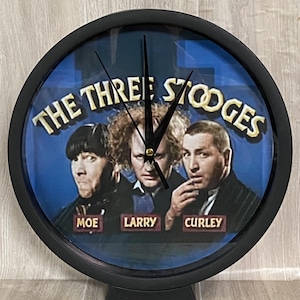 May include: A black, round clock featuring the Three Stooges. The clock face has a blue background with the faces of Moe, Larry, and Curley. The words "The Three Stooges" are in gold lettering above the faces. The clock has black hands and a black frame.