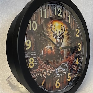 May include: A round black wall clock featuring a forest scene with a deer. The clock face has large gold numerals and hands. The background shows a deer in a forest with a sunset. The clock has a clear plastic cover.