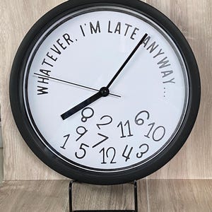 May include: A round black and white clock with the text "WHATEVER, I'M LATE ANYWAY...". The clock face has black numbers and hands. The clock is displayed on a black metal stand.