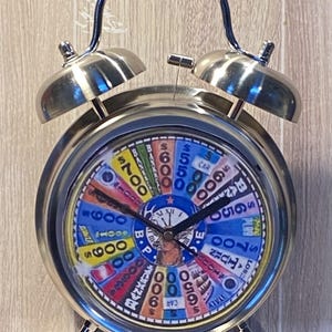 May include: A silver-colored alarm clock with a colorful face resembling a game show wheel. The clock has a curved handle and two bells on top. The face features numbers, text, and various colors.
