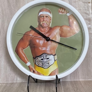 May include: A white-framed wall clock featuring a muscular man in yellow trunks, a championship belt, and a white headband with the word "Hulkster." The clock has black hands and a black stand. The background is a light green color.