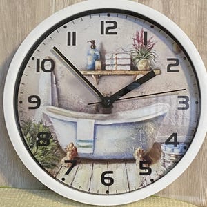 May include: A round wall clock with a white frame and black numbers. The clock face features a printed illustration of a clawfoot bathtub, a wooden shelf with stacked towels, a soap dispenser, and a potted plant on a wooden floor.