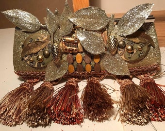 May include: A gold clutch purse with a glittery finish and a design of leaves and other embellishments. The purse has a fringe trim with tassels in shades of brown and orange.