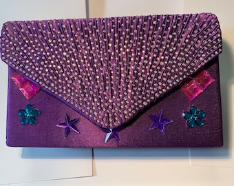 May include: A purple satin clutch purse with a sparkly design. The purse has a flap closure and is decorated with rhinestones, stars, and flowers.