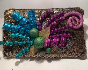 May include: A gold sequined clutch bag with a decorative design featuring blue, green, and purple embellishments. The design includes a swirl, beads, and ribbons.