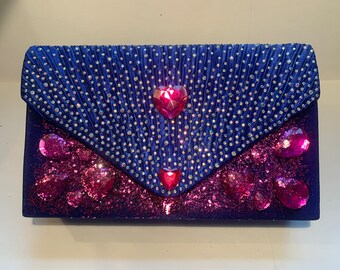May include: A blue velvet clutch purse with pink glitter and rhinestones. The purse has a heart-shaped rhinestone in the center.