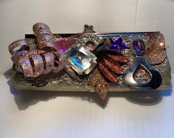 May include: A glittery silver clutch with a silver clasp, adorned with sparkling embellishments. Features a large clear crystal, pink and purple accents, and a silver teardrop-shaped charm. Decorative and festive.