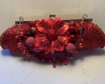 May include: A red glitter clutch purse with a silver chain strap and a large red flower embellishment. The flower has a pink center and is surrounded by red beads.
