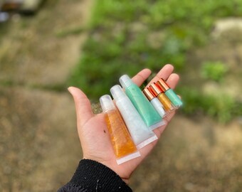May include: A hand holding five clear lip gloss tubes with gold and copper lids. The lip glosses are in shades of green, white, and gold.