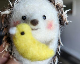 May include: A white needle felted hedgehog with brown eyes and a pink blush holding a yellow needle felted crescent moon.