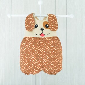 May include: A brown and white polka dot baby onesie with a dog face on the front. The onesie has brown ears and a brown nose. The onesie is hanging on a white hanger.