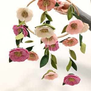 May include: A hanging mobile featuring multiple felt flowers in shades of pink, cream, and white, each with a textured gold centre. The flowers are attached to a circular wooden hoop by white strings, interspersed with green felt leaves.