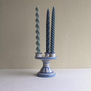 May include: A ceramic candle holder with a blue and white striped pattern holds three tall, twisted taper candles. The candles are in varying shades of blue, ranging from a light powder blue to a deeper navy, each featuring a spiral texture.