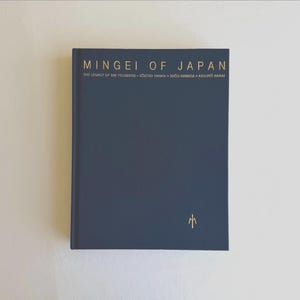 May include: A dark blue hardcover book titled MINGEI OF JAPAN: THE LEGACY OF THE FOUNDERS - SŌETSU YANAGI • SHŌJI HAMADA • KANJIRŌ KAWAI. The front cover features gold lettering and a small gold symbol near the bottom right corner.