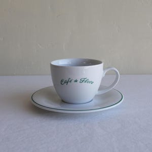 Op de afbeelding: Witte keramische theekop en schotel set. De kop heeft een oor en de woorden "Café de Flore" in groene letters. De schotel heeft een groene rand. De set staat op een wit oppervlak.