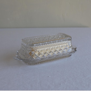 May include: A clear glass butter dish with a rectangular shape and a diamond pattern. The lid and base have decorative edges. The dish is sitting on a white surface.