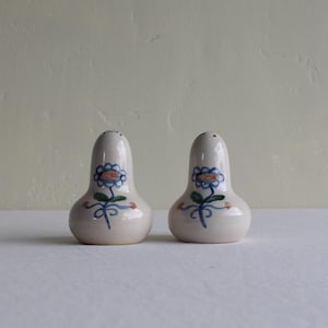 Puede incluir: Un par de saleros y pimenteros de cerámica. Los saleros son de color blanco roto, con una base bulbosa y una parte superior estrecha. Cada salero está decorado con un diseño floral azul y naranja pintado a mano y hojas verdes.