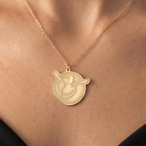 Può includere: Una collana con pendente dorato con un pendente circolare con un disegno inciso. Il pendente pende da una delicata catena d'oro. La collana è indossata sopra un top nero.