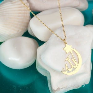 Peut inclure: Un collier dor&eacute; compos&eacute; d'une cha&icirc;ne fine et d'un pendentif en forme de croissant de lune surmont&eacute; d'une &eacute;toile. Le pendentif pr&eacute;sente la calligraphie arabe du mot Allah int&eacute;gr&eacute;e au design du croissant de lune, formant un bijou coh&eacute;rent.