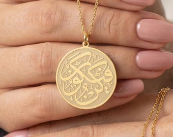 May include: A gold-colored necklace with a circular pendant featuring ornate calligraphy. The pendant hangs from a delicate chain. The necklace is held up by a person with pink nail polish.