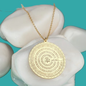 Pu&ograve; includere: Una collana con ciondolo circolare color oro caratterizzata da un'intricata calligrafia araba incisa in cerchi concentrici. Il ciondolo pende da una delicata catenina color oro e presenta un motivo dettagliato sul bordo.