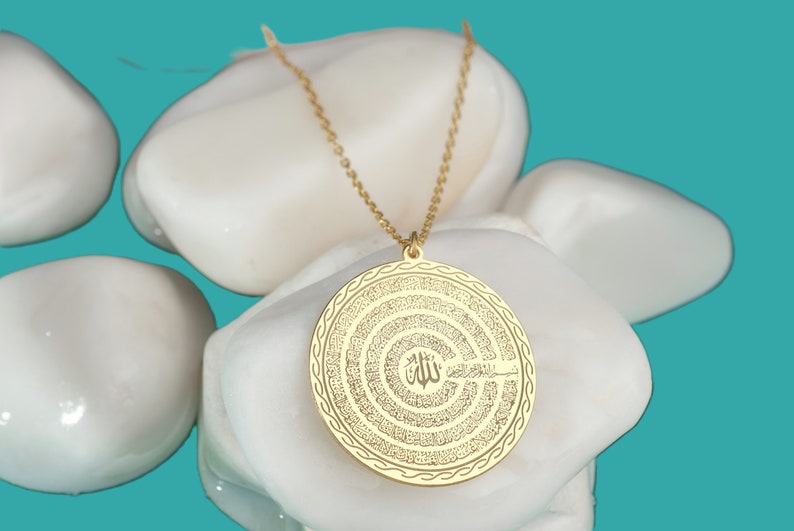 Pu&ograve; includere: Una collana con ciondolo circolare color oro caratterizzata da un'intricata calligrafia araba incisa in cerchi concentrici. Il ciondolo pende da una delicata catenina color oro e presenta un motivo dettagliato sul bordo.