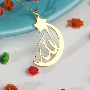 Peut inclure: Un collier pendentif en m&eacute;tal dor&eacute; repr&eacute;sentant un croissant de lune int&eacute;gr&eacute; &agrave; une &eacute;toile &agrave; cinq branches sur le dessus. Le centre du croissant de lune pr&eacute;sente une calligraphie arabe. Le pendentif est suspendu &agrave; une fine cha&icirc;ne en m&eacute;tal dor&eacute;.