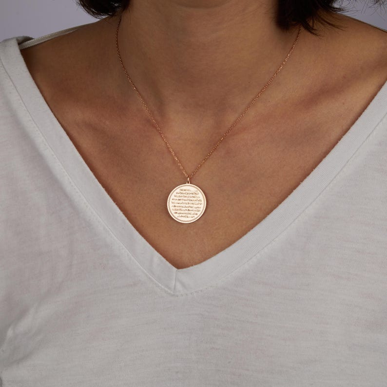 May include: A rose gold necklace with a circular pendant. The pendant features engraved text and hangs from a delicate chain. The necklace is worn against a white v-neck shirt.