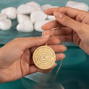Pu&ograve; includere: Una collana con ciondolo circolare color oro caratterizzata da intricati motivi incisi concentrici e calligrafia araba centrale. Il ciondolo &egrave; sospeso a una delicata catenina dorata, mostrando un design superficiale dettagliato e strutturato.