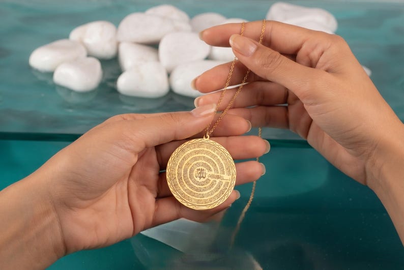 Pu&ograve; includere: Una collana con ciondolo circolare color oro caratterizzata da intricati motivi incisi concentrici e calligrafia araba centrale. Il ciondolo &egrave; sospeso a una delicata catenina dorata, mostrando un design superficiale dettagliato e strutturato.