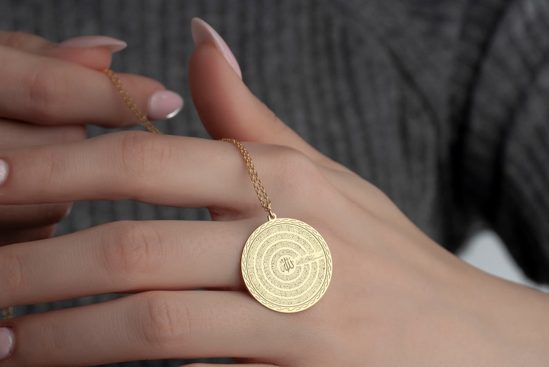 Pu&ograve; includere: Una collana con ciondolo circolare color oro caratterizzata da un intricato motivo a spirale concentrica inciso sulla superficie. Il centro del disco mostra un piccolo e distinto simbolo calligrafico arabo, sospeso a una delicata catenina dorata.