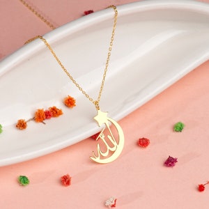 Peut inclure: Un collier de couleur dor&eacute;e avec un pendentif en forme de croissant de lune et d'&eacute;toile, int&eacute;grant une calligraphie arabe dans le design de la lune. Le pendentif est suspendu &agrave; une fine cha&icirc;ne dor&eacute;e, pr&eacute;sentant une finition m&eacute;tallique polie.