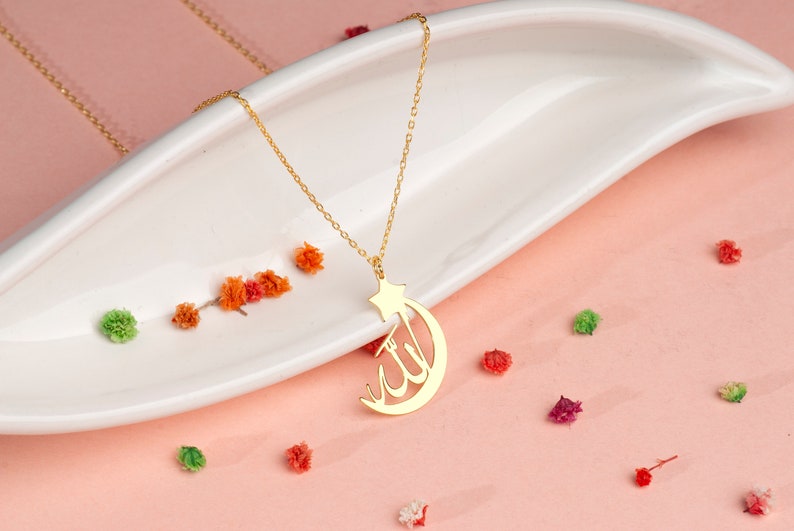 Peut inclure: Un collier de couleur dor&eacute;e avec un pendentif en forme de croissant de lune et d'&eacute;toile, int&eacute;grant une calligraphie arabe dans le design de la lune. Le pendentif est suspendu &agrave; une fine cha&icirc;ne dor&eacute;e, pr&eacute;sentant une finition m&eacute;tallique polie.