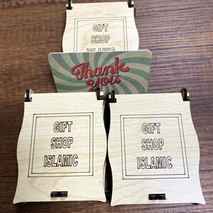 Pu&ograve; includere: Tre scatole in legno chiaro con lati curvi, ognuna con un bordo quadrato e il testo GIFT SHOP ISLAMIC inciso sulla parte anteriore. &Egrave; inclusa una carta rettangolare con un motivo a raggiera verde e bianco e il testo rosso Thank you.
