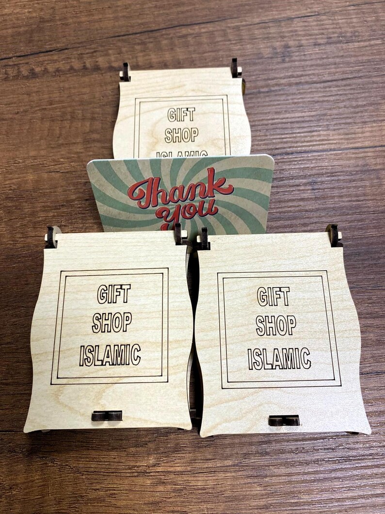 Pu&ograve; includere: Tre scatole in legno chiaro con lati curvi, ognuna con un bordo quadrato e il testo GIFT SHOP ISLAMIC inciso sulla parte anteriore. &Egrave; inclusa una carta rettangolare con un motivo a raggiera verde e bianco e il testo rosso Thank you.