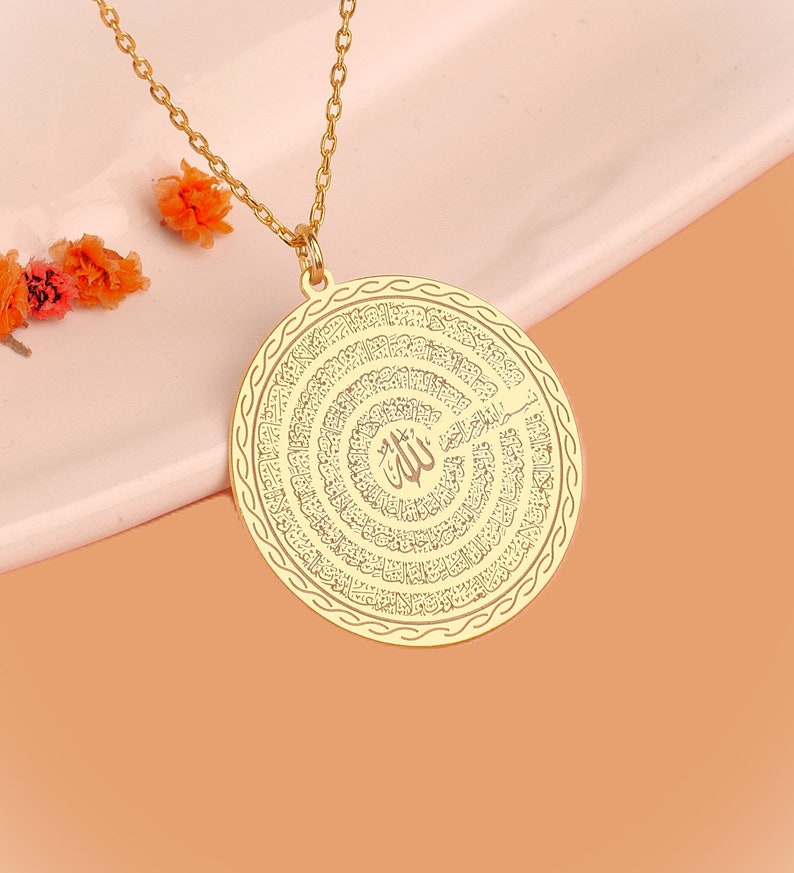 Pu&ograve; includere: Una collana con ciondolo circolare color oro caratterizzata da un'intricata calligrafia araba incisa a spirale. Il centro del disco mostra la parola Allah, circondata da linee di testo concentriche e da un bordo decorativo lungo il perimetro.