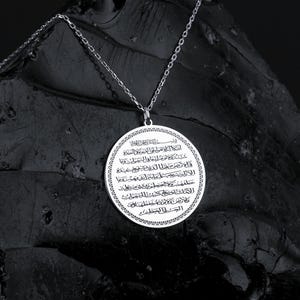 May include: A silver-colored pendant necklace with a circular pendant. The pendant features intricate Arabic script and is suspended from a delicate chain. The necklace is displayed against a dark, textured background.