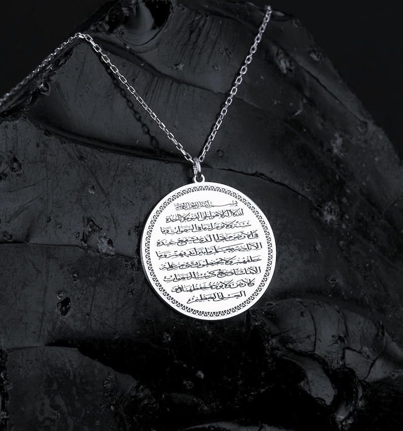 May include: A silver-colored pendant necklace with a circular pendant. The pendant features intricate Arabic script and is suspended from a delicate chain. The necklace is displayed against a dark, textured background.