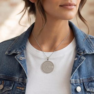 Pu&ograve; includere: Una collana con ciondolo circolare color argento caratterizzata da una serie di cerchi concentrici e un piccolo motivo a stella centrale. Il ciondolo pende da una sottile catenina argentata su una t-shirt bianca sotto una giacca di jeans.