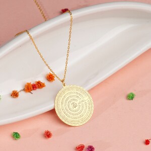 Pu&ograve; includere: Una collana con ciondolo circolare color oro caratterizzata da un intricato motivo testurizzato con calligrafia araba al centro. Il ciondolo pende da una delicata catenina color oro, mostrando un bordo lucido e un dettagliato design inciso.
