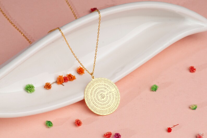 Pu&ograve; includere: Una collana con ciondolo circolare color oro caratterizzata da un intricato motivo testurizzato con calligrafia araba al centro. Il ciondolo pende da una delicata catenina color oro, mostrando un bordo lucido e un dettagliato design inciso.